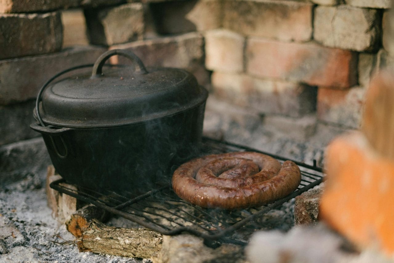 Cast iron pot and sausage on grill outdoors in rustic setup.