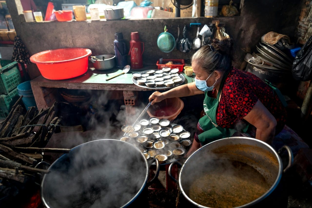 Elderly woman in a mask preparing traditional dishes in a rustic village kitchen.