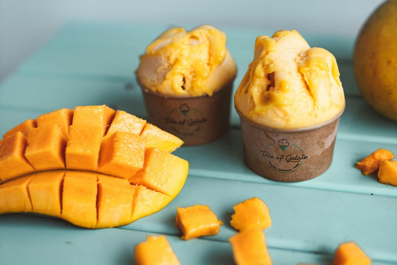 Mango gelato scoops in cups with fresh mango slices on a turquoise table.